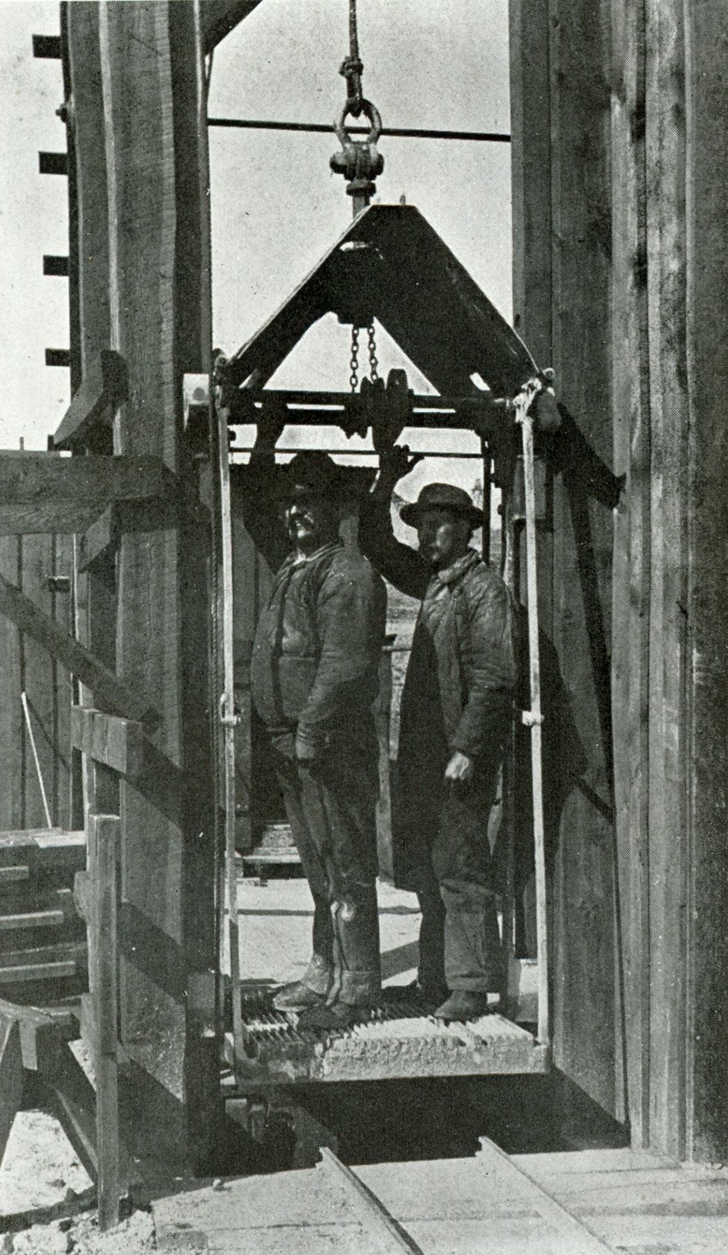 Two Butte miners standing in an open cage as it is lowered into a timbered mine shaft, about 1900